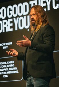 Speaker with long hair and beard in a black blazer giving a talk on stage, large white text behind him.
