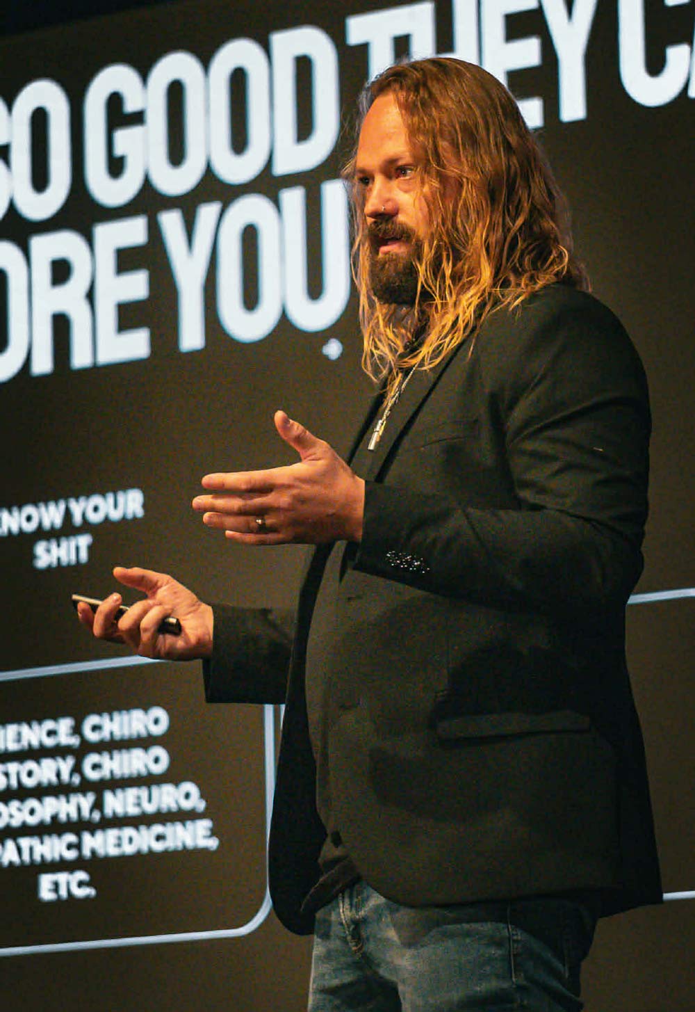 Speaker with long hair and beard in a black blazer giving a talk on stage, large white text behind him.