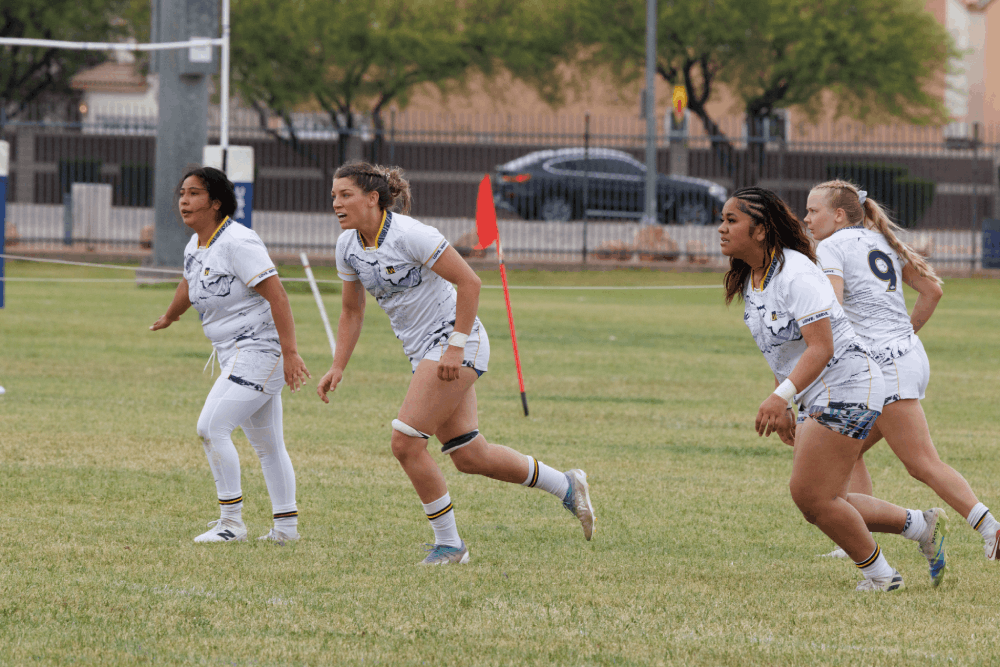 Four female rugby players in white uniforms sprint across a grassy field during a match, with a red flag marker in the background.