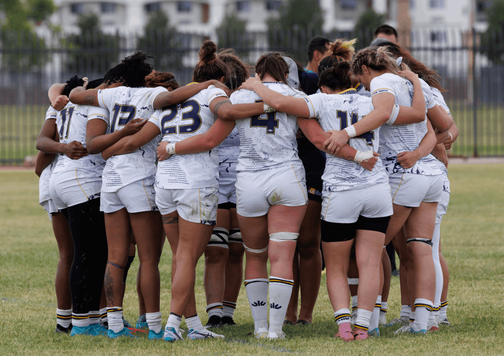 Female rugby players wearing white uniforms form a tight circle in a team huddle on a grassy field before a game, with arms around each other.