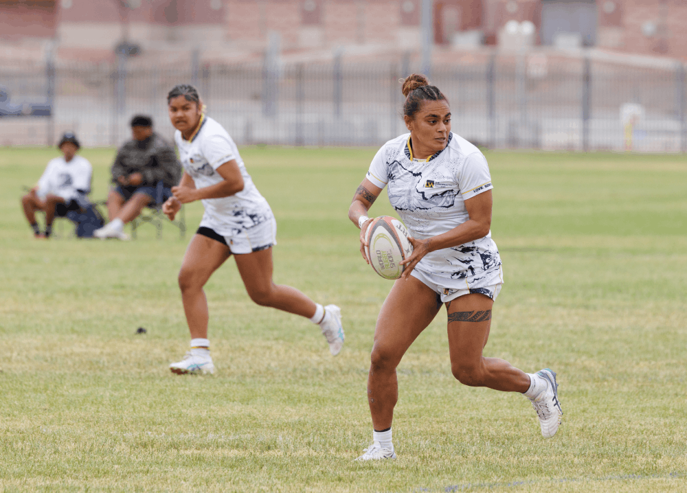 Female rugby player in a white uniform runs with a ball on a grassy field, with teammates in the background.