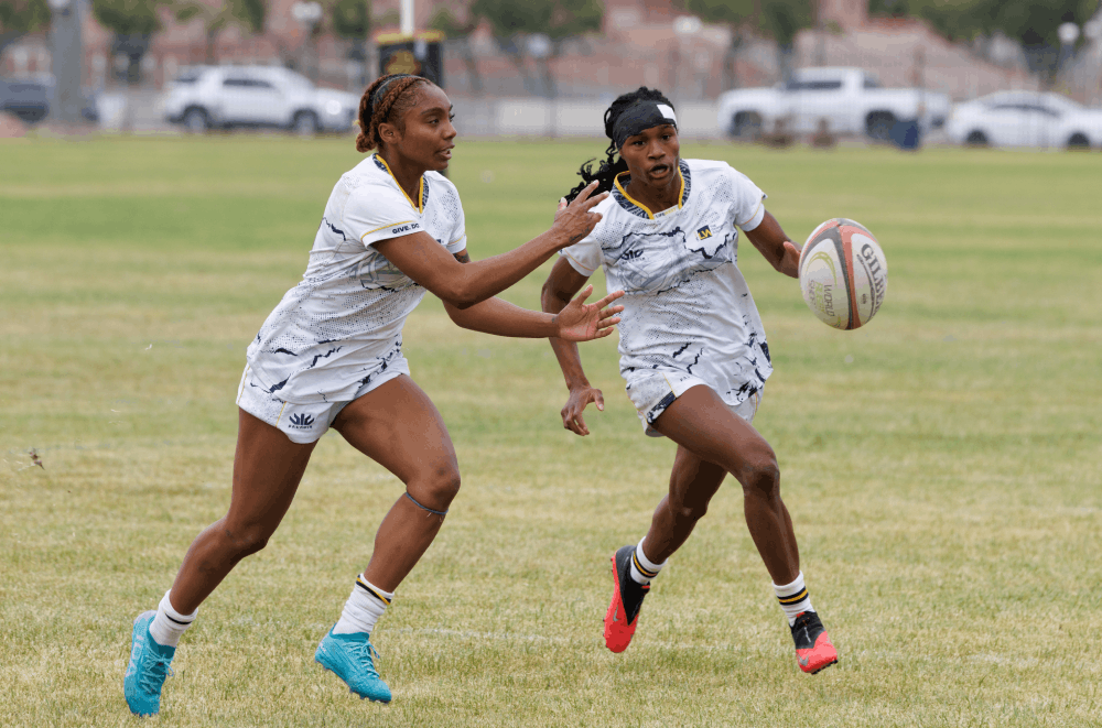 Two female rugby players in white uniforms sprint on a grassy field, one holding the ball as they run toward the try area