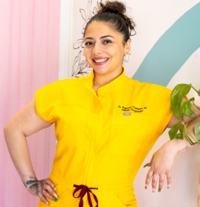 Woman in a bright yellow jumpsuit posing with one hand on her hip, smiling at the camera in a colorful room with pink paneling and teal arc shapes.