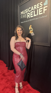 Woman in a red strapless dress posing on a red carpet beside a 'Musicares Fire Relief' backdrop. She holds a black clutch and smiles at the camera.