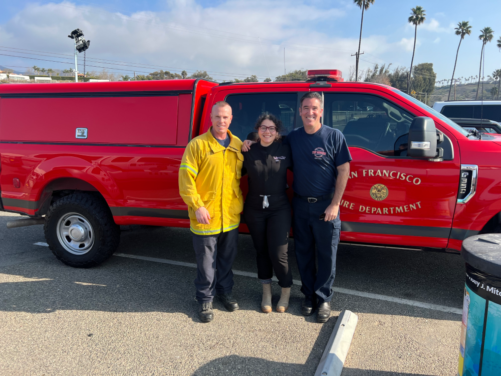 Three people stand arm in arm in front of a red San Francisco Fire Department vehicle, smiling outdoors with palm trees in the background.