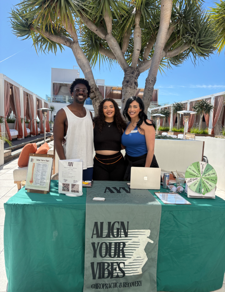 Three smiling people stand behind a teal table at an outdoor resort promo booth for Align Your Vibes Chiropractic & Recovery.