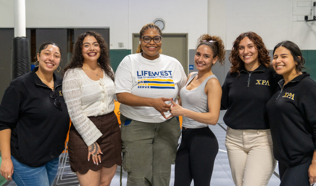 Six women stand together in a gym, smiling at the camera, wearing casual clothes and branded apparel.