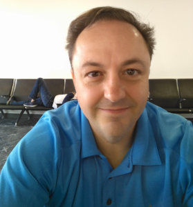 Man in blue polo taking a selfie in a waiting area, with a person lying on a bench in the background.