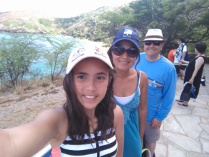 Girl taking a selfie with two adults at a seaside overlook; turquoise water and green cliffs in the background.