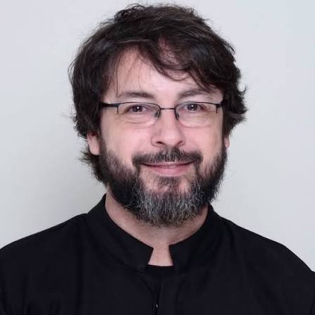 Portrait of a man with dark hair and a beard wearing glasses, smiling at the camera in a black shirt.