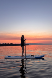 Person standing on a paddleboard at sunset on a calm lake, holding a paddle.
