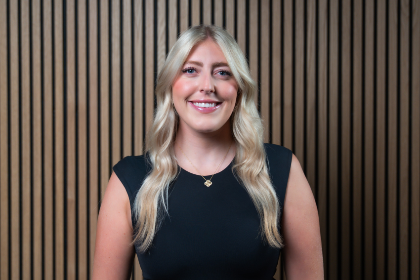 Smiling woman with long blonde hair wearing a black sleeveless top, standing against a vertical wooden slat wall.